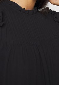 Haut noir avec texture plissée, encolure volantée et manches courtes à volants. Tissu léger, design simple sans motifs visibles.