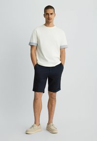 Camiseta de hombre de color crema con puños a rayas, combinada con pantalones cortos de un azul marino oscuro. Calzado beige complementa el atuendo, todo contra un fondo claro.