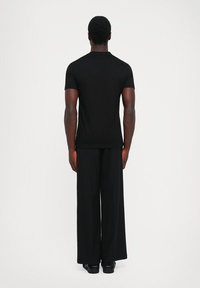 Bărbat stând de spate, îmbrăcat cu un tricou simplu negru cu mânecă scurtă, pantaloni negri largi și adidași negri cu logo-ul Versace.