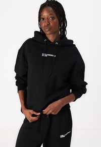 Musthave musta kapuutsiga sviiter, millel on soonikkoes varrukad ja nöör, ning mille rinnal on valge logo tekst. Komplekti kuuluvad ka sobivad mustad spordipüksid, millel on samuti logo.