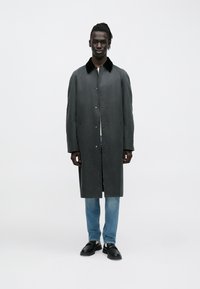 Lang grå frakke med sort krave, udstyret med knapper og sidelommer, båret over lyseblå jeans og sorte sko.