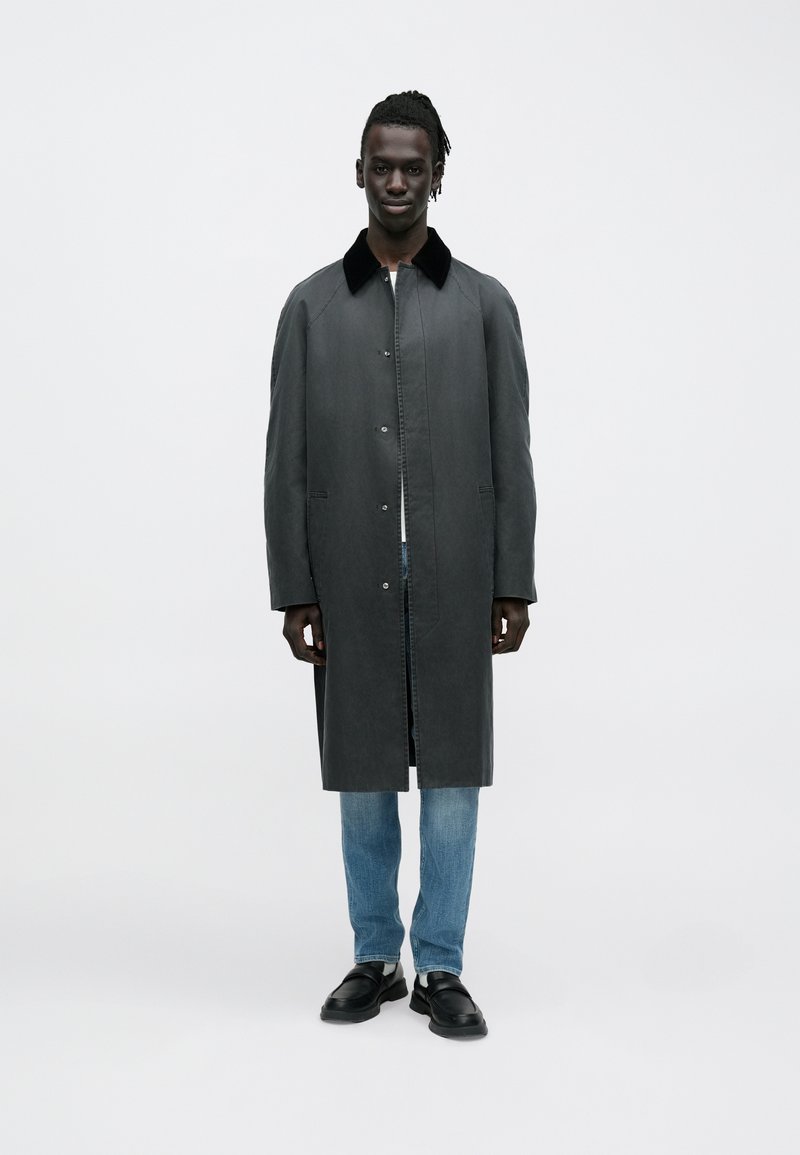 Lang grå frakke med sort krave, udstyret med knapper og sidelommer, båret over lyseblå jeans og sorte sko.