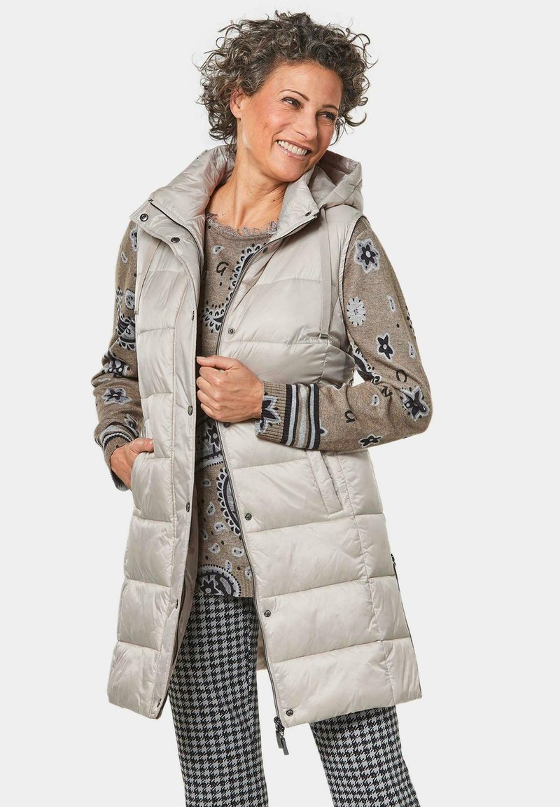 Hellbeige gepolsterte Weste mit Kapuze, ausgestattet mit Druckknöpfen, Seitentaschen und einer schimmernden Textur, über einem gemusterten Pullover und karierten Hosen getragen.