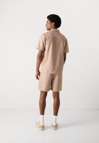 Conjunto de color beig de manga corta con un patrón sutil, que incluye una camisa con cuello y pantalones cortos a juego. El material parece ligero y texturizado.