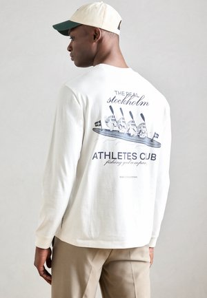 Biele tričko s dlhým rukávom a potlačou na zadnej strane; v kombinácii s béžovými nohavicami. Tričko má rebrované manžety a voľný strih.