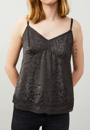 Femme portant un caraco noir à motif floral avec fines bretelles et un jean foncé, avec des tatouages de symboles du zodiaque sur le haut du bras gauche.