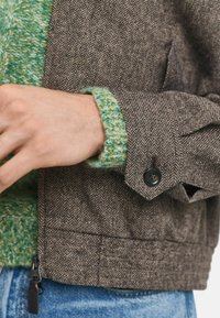 Bruine jas met een visgraatpatroon, voorzien van een teal gebreide manchette en zwarte knoopdetails. Heeft een gestructureerd design en een textuurafwerking.
