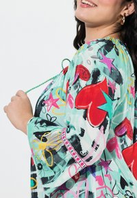 Blouse légère et colorée de couleur menthe avec des motifs de cœurs et d'étoiles. Elle présente un col rond, des manches évasées et une garniture décorative.