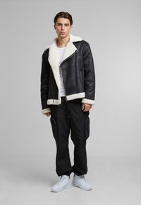Chaqueta de borreguillo negra con forro de piel blanca, cierre de cremallera, solapas anchas y puños en contraste. Combinada con pantalones cargo negros y zapatillas blancas.