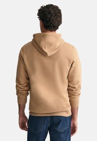 Felpa con cappuccio beige, realizzata in tessuto morbido con polsini a costine, tasca a marsupio e vestibilità comoda. Indossata con jeans blu.