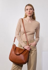 Sac en cuir marron avec une poignée tressée, en forme de goutte d'eau, et une bandoulière amovible. Porté par-dessus un haut beige clair et un pantalon crème.