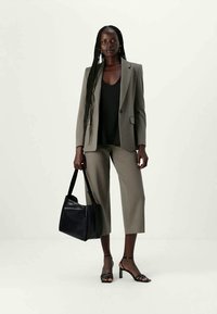 Mujer alta con trenzas largas que lleva un blazer topo y pantalones cortos, una blusa negra, sandalias de tacón negro y lleva un gran bolso negro.