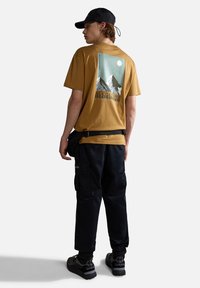 Camiseta gráfica marrón con un diseño de montaña, combinada con pantalones cargo negros y zapatillas negras con suelas texturizadas. La gorra es negra.