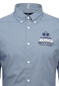 Light blue button-up shirt with a pointed collar, featuring white buttons and embroidered navy text and logo for "La Martina Argentin Polo Team."