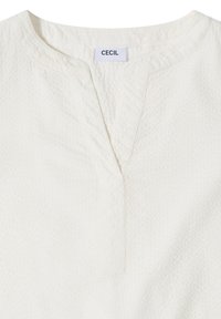 Chemise blanche texturée avec un col en V et une étiquette de marque "CECIL" cousue à l'intérieur de l'encolure.