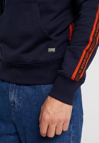 Primo piano di una persona che indossa un hoodie blu scuro con strisce rosse G-STAR ORIGINALS sulle maniche e jeans in denim blu, con la mano rilassata lungo il fianco.