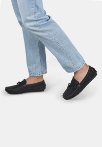 Melni ādas loafers ar plakanu zoli, ar kontrastējošām šuvēm, šnoru detaļu un gumijas akcentiem aizmugurē. Valkāti ar gaiši ziliem džinsiem.