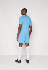 Blauwe trui en shorts van lichtgewicht materiaal. Voorzien van een gouden logo op de schouder. Witte sokken en zwarte voetbalschoenen.