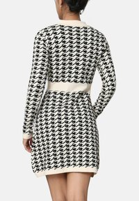 Femme portant une robe à manches longues en damier noir et blanc avec une ceinture crème, les mains sur les hanches, de dos face au spectateur.