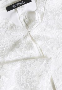 Robe en dentelle blanche avec motifs floraux, doublure intérieure lisse et fermeture éclair discrète dans le dos. L'étiquette indique "SWING".