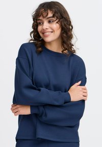 Námořnická modrá mikina z měkké, texturované látky s volnějším střihem, kulatým výstřihem a mírně spuštěnými rameny. Minimální designové detaily.