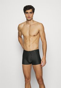 Zwart gestreepte boxershorts met een aansluitend ontwerp en een gladde textuur, met een logo op de tailleband. Model staat tegen een effen achtergrond.