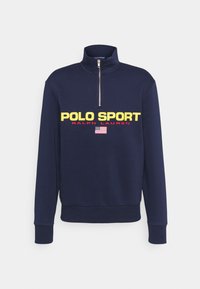 Marineblaue Sweatshirt mit hohem Kragen, halber Reißverschluss, mit "POLO SPORT" in gelber und roter Schrift, darunter ein Detail der amerikanischen Flagge.