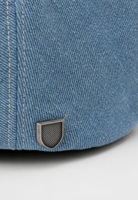 Világoskék farmer sapka texturált felülettel. Fémes pajzs logóval rendelkezik csíkozott mintával és kontrasztos varrással.