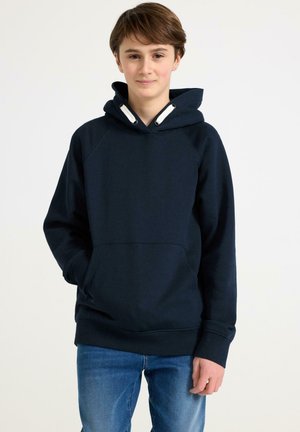 Teenagedreng står med hænderne i lommerne, iført en mørk blå hættetrøje og blå jeans mod en ensfarvet lys baggrund.