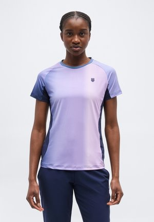 Žena v krátkym rukávom športovom tričku fialovej a modrej farby a tmavomodrých nohaviciach stojaca pred jednoduchým bielym pozadím.