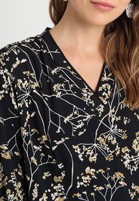 Mustassa kankaasta valmistettu paita, jossa on vaaleita kukkakuoseja valkoisena ja kultaisena, V-aukkoinen kaula-aukko ja teksturoitu pinta.