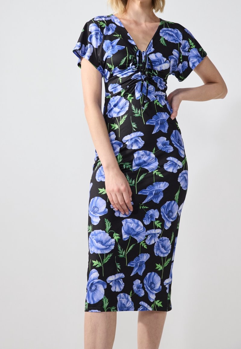 Femme portant une robe noire mi-longue avec des imprimés floraux bleus et des manches courtes, debout avec une main sur la hanche.