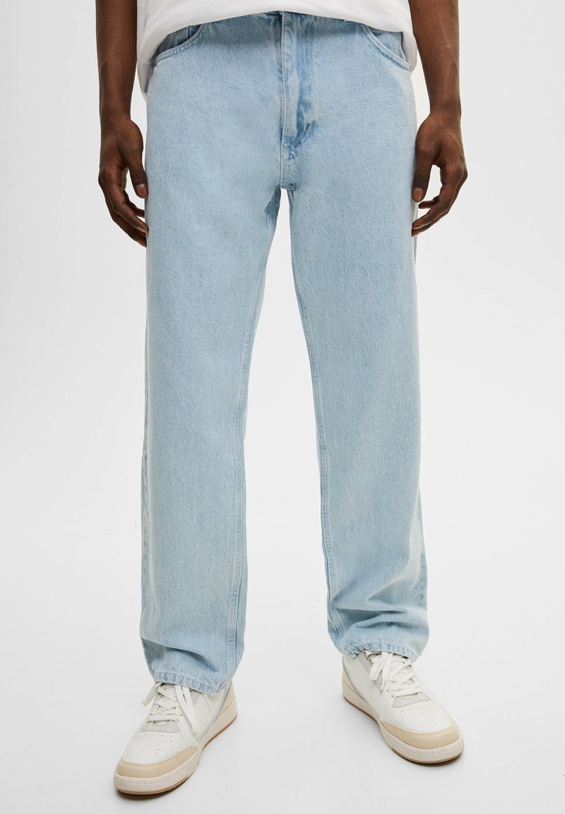 Jeans de mezclilla azul claro con corte recto. Presentan un bolsillo frontal y un cierre de botón, combinados con zapatillas blancas para un look casual.