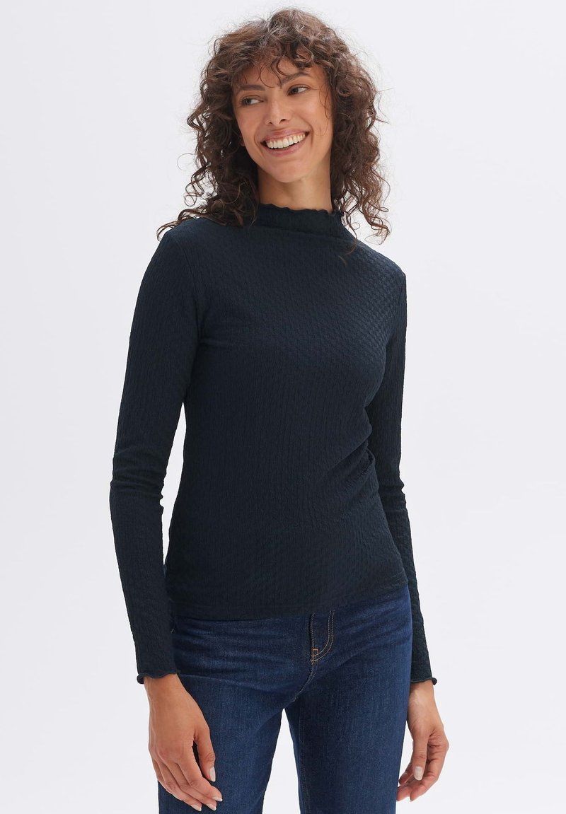 Schwarzes Langarmshirt mit einem strukturierten Muster und einem rüschenbesetzten hohen Ausschnitt, kombiniert mit dunkelblauen Jeans. Glattes Material und taillierter Schnitt.