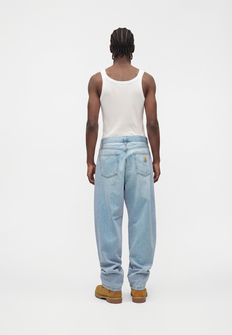 Man with braided hair wearing a white sleeveless shirt, light blue loose jeans, and brown boots standing facing away on a plain white background.