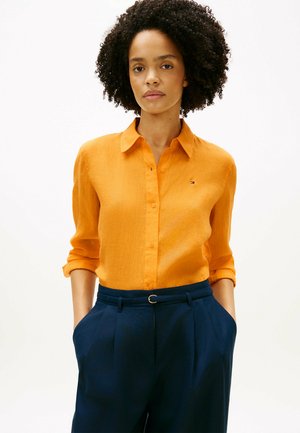 Mujer con cabello rizado que lleva una camisa naranja con las mangas remangadas y pantalones azul marino con cinturón, manos en los bolsillos, fondo neutro.