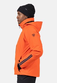 Ljusröd vattentät jacka med huva, svarta detaljer och två sidofickor; har en logotyp på överarmen och randiga detaljer.