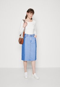 Frau mit Brille, weißem Langarmoberteil, zweifarbigem blauen Jeansrock, weißen Slippers und einer braunen Umhängetasche.