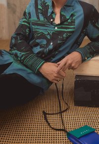 Shirt met groen en zwart grafisch ontwerp, knopen en kraag; gecombineerd met een leren tas in blauw en groen met een schouderriem.