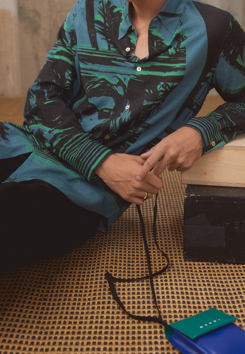 Košile s grafickým designem v zelené a černé barvě, s knoflíky a límečkem; spárovaná s koženou taškou v modré a zelené barvě s popruhem.