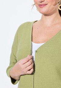 Ljusgrön kofta med strukturerad väv, med V-hals, en enkel knappknäppning och trekvartsärmar, bärs över en vit topp.