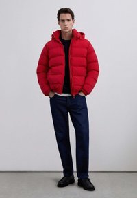 Joven con una chaqueta acolchada roja, jeans oscuros y zapatos negros de pie contra un fondo gris liso con las manos en los bolsillos.