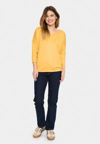 Gul, överdimensionerad stickad tröja med 3/4 ärmar, kombinerad med mörkblå jeans och beiga sneakers med gröna ränder. Enkelt mönster och mjuk textur.