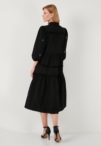 Rochie neagră midi cu sleeve puff, volane și broderie florală albastră subtilă. Prezintă un corset ajustat și o fustă evazată. Sandale cu toc negre.
