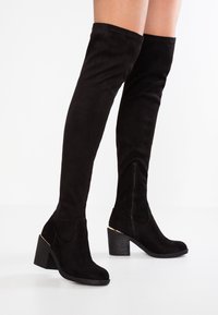 Paire de cuissardes en daim noir avec talons carrés et fermetures éclair latérales portées sur des jambes à teint clair sur fond blanc.