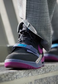 Gros plan d'une basket Nike grise et violette avec une bride à logo bleu portée sous un pantalon gris à carreaux, debout sur un rebord en pierre.