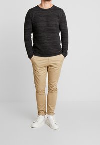 Maglione a coste nero con scollatura rotonda, abbinato a chinos beige skinny e sneakers bianche, in piedi contro uno sfondo neutro.