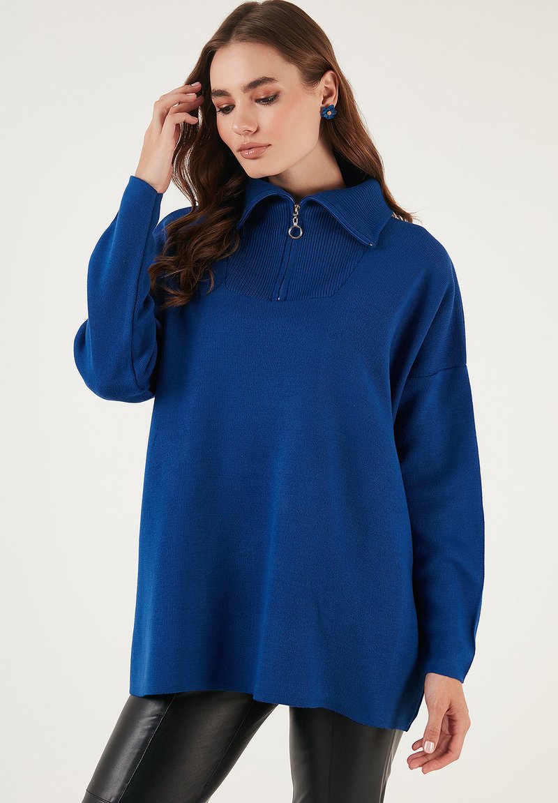 Pulover albastru cu guler tip fermoar, detalii cu nervuri și mâneci lungi. Purtați peste pantaloni negri, cu textură netedă.