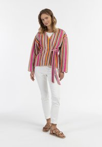 Giacca a maglia multicolore a strisce con maniche lunghe e cintura da annodare, abbinata a pantaloni bianchi e sandali marroni con cinturini.