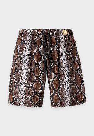 Muskelmand står barfodet med den ene hånd i lommen iført brune og beige shorts med slangeprint på en ensfarvet grå baggrund.